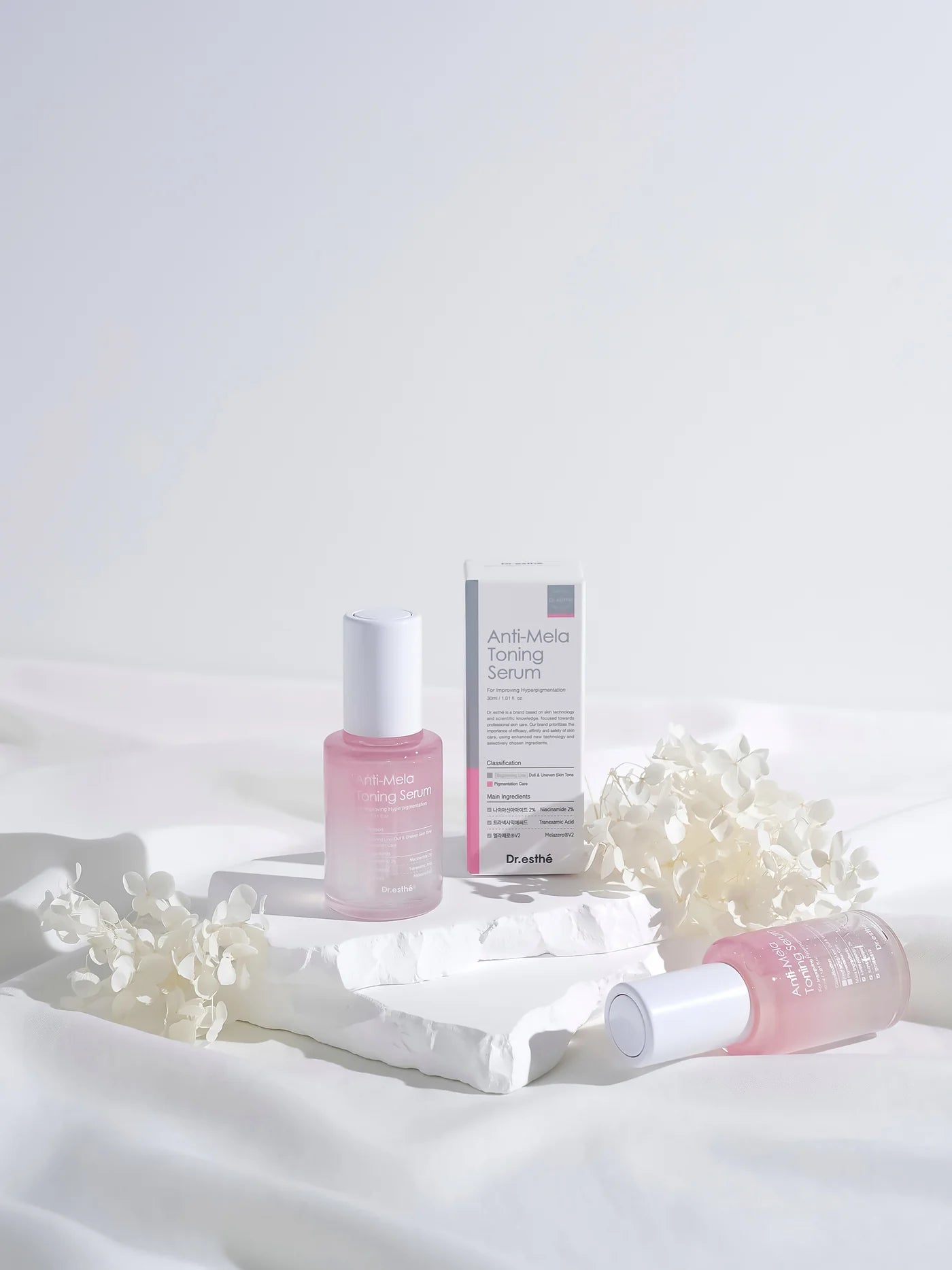Dr. esthé Anti-Mela Toning Serum bottle and box displayed on white stone with dried flowers, highlighting Melazero V2, Niacinamide 2%, and dermatological testing.