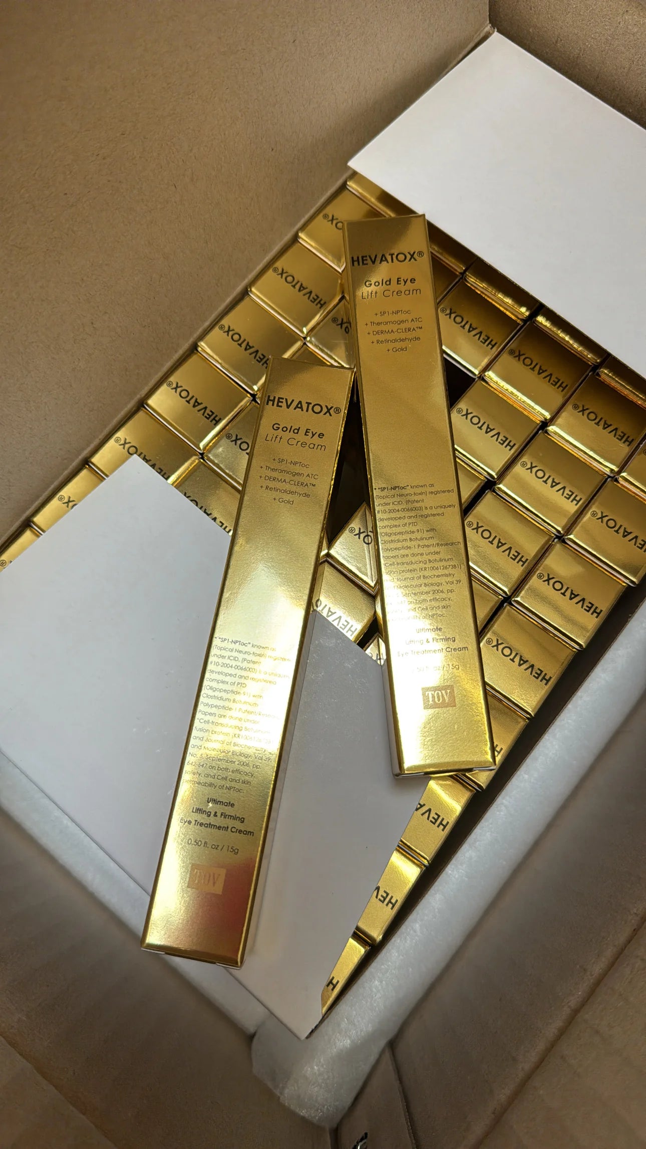 Stack of Hevatox Gold Eye Lift Cream retail boxes in a shipping carton, with two boxes propped up showing ingredient callouts and patent research citations on the side panel.
