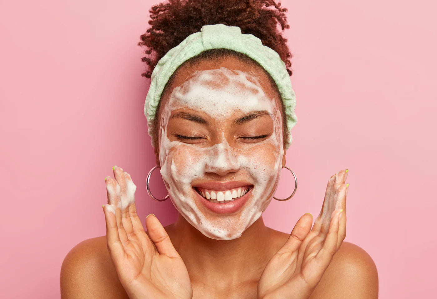 Woman with a green headband smiling with a foaming cleanser applied to her face against a pink background.
